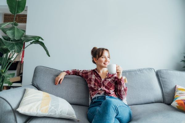 Young smiling woman sitting on sofa and looking side up while drinking coffee or tea